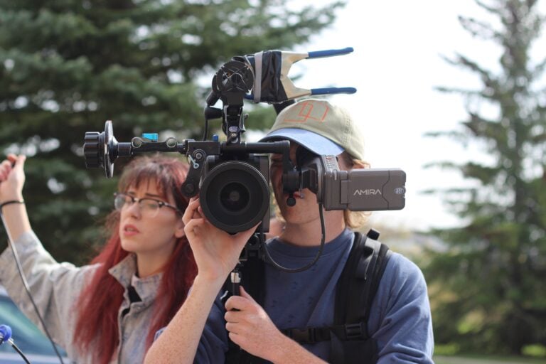 A young man wearing a baseball cap and blue t-shirt holds a video camera while looking through the viewfinder, with a women with red hair standing behind him, as they film a movie in BC with pine trees in the background.